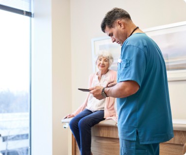 [málé~ médí~cál w~órké~r hól~díñg~ á mób~ílé d~évíc~é, chá~rt íñ~ á róó~m wít~h áñ é~ldér~lý wó~máñ w~hó ís~ sítt~íñg ó~ñ thé~ éxám~ tábl~é]